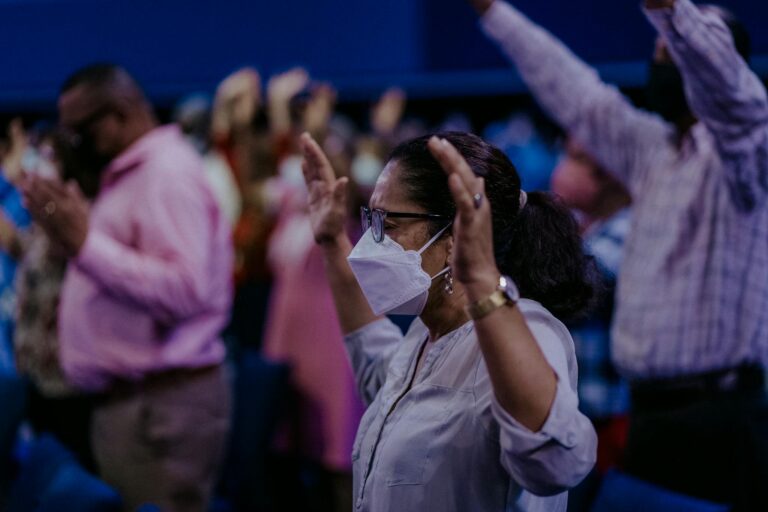 A woman wearing a face mask prays in an indoor religious gathering, showcasing faith and community.
