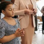 A young girl in a floral dress silently prays during a community gathering indoors.