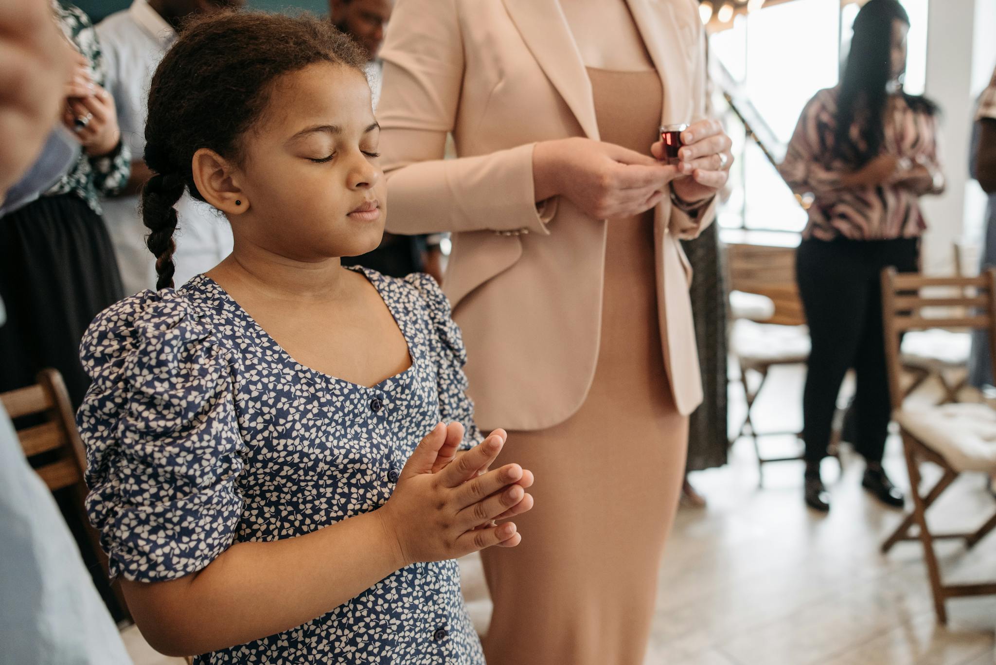 A young girl in a floral dress silently prays during a community gathering indoors.