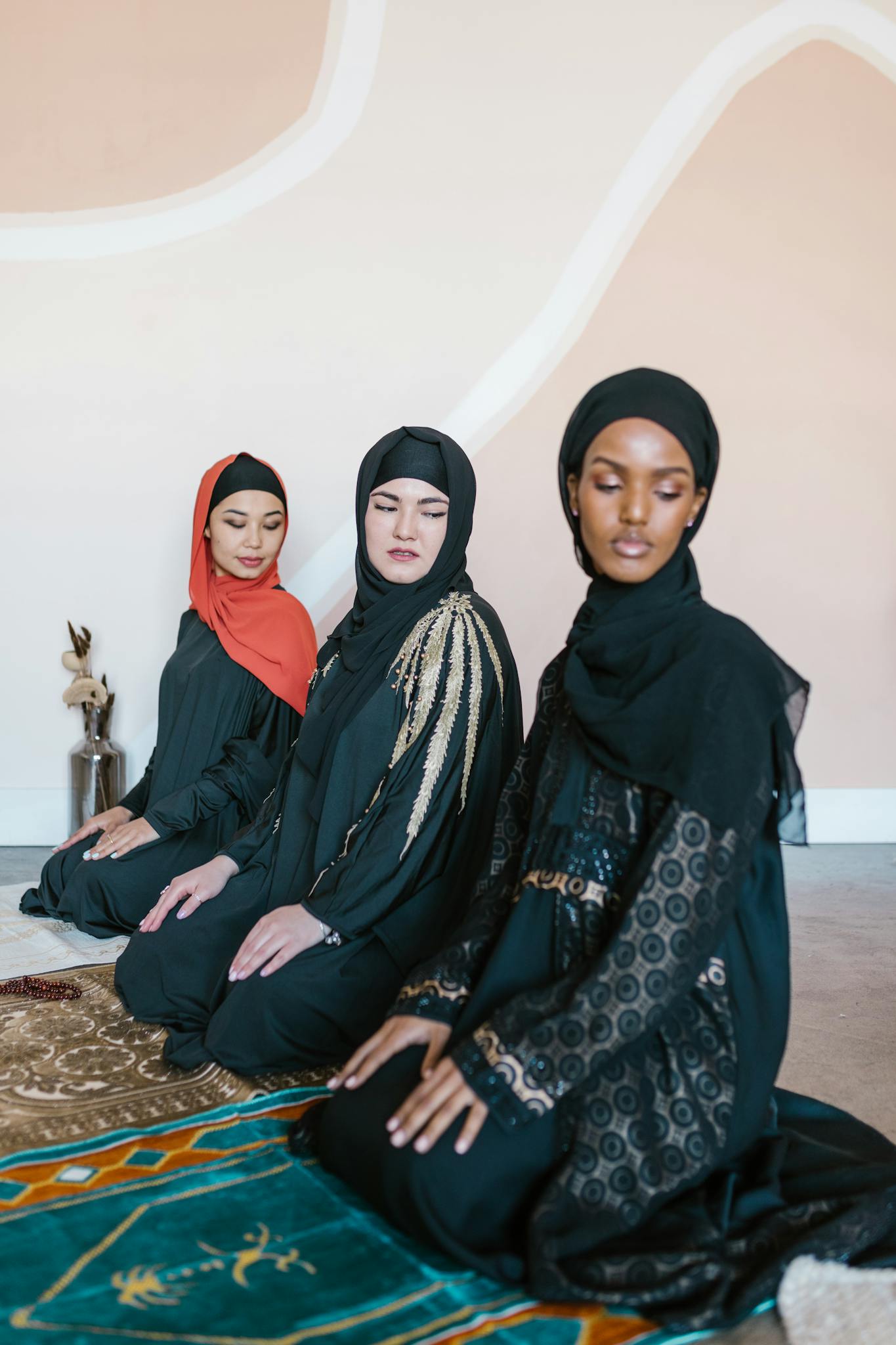 Three women in hijabs kneeling on prayer rugs indoors, embracing cultural and religious traditions.